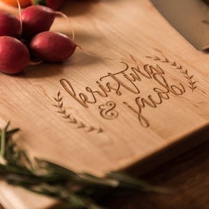 Peut inclure: Une planche &agrave; d&eacute;couper en bois avec le texte "Kristina & Jacob" grav&eacute; dessus. La planche est de couleur brun clair et pr&eacute;sente un grain de bois naturel. Il y a quelques radis et des brins de romarin sur la planche.