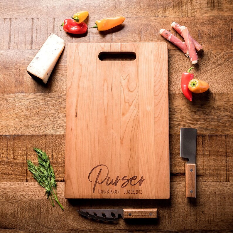 May include: A personalized wooden cutting board with the text "Purser Brian & Karen June 23, 2012" engraved on it. The board is light brown and has a handle.
