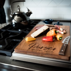 May include: A wooden cutting board with the text "Purser Brian & Karen June 23, 2012" engraved on it. The board is on a black and silver stovetop and has a knife, cheese, salami, and peppers on it.