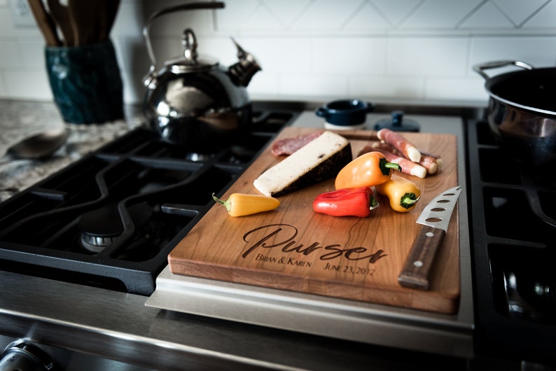 May include: A wooden cutting board with the text "Purser Brian & Karen June 23, 2012" engraved on it. The board is on a black and silver stovetop and has a knife, cheese, salami, and peppers on it.