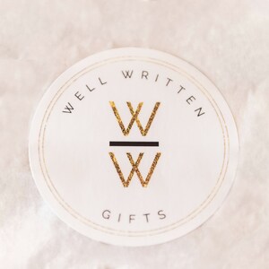 May include: A white round sticker with a gold double-lined border. The text "WELL WRITTEN" is written in black around the top of the sticker. The text "GIFTS" is written in black at the bottom of the sticker. A gold "W" is stacked on top of another gold "W" with a black line separating the two letters.