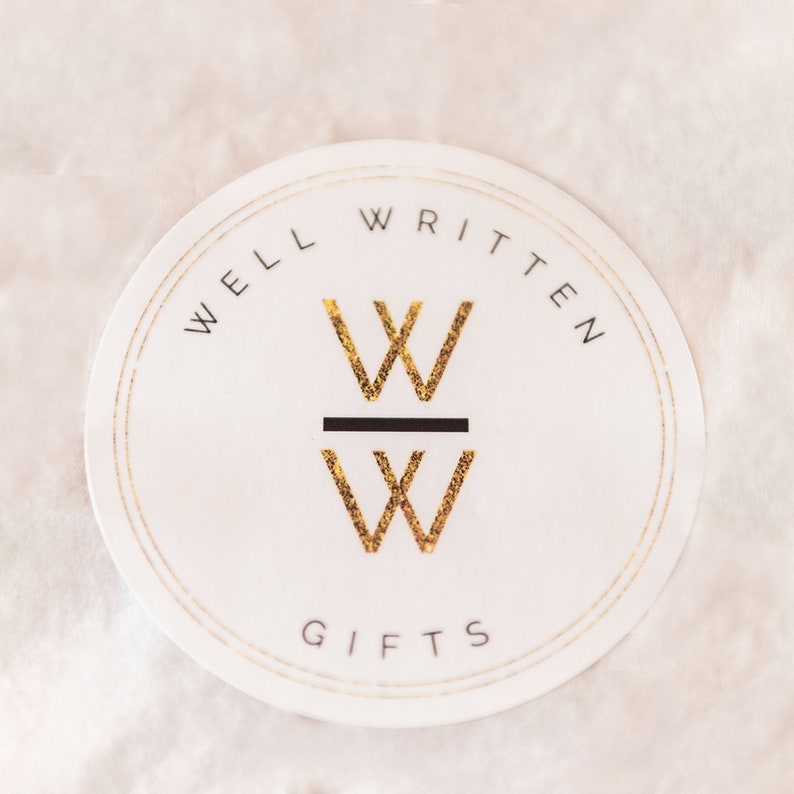 May include: A white round sticker with a gold double-lined border. The text "WELL WRITTEN" is written in black around the top of the sticker. The text "GIFTS" is written in black at the bottom of the sticker. A gold "W" is stacked on top of another gold "W" with a black line separating the two letters.