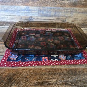 May include: A clear glass baking dish sits atop a patriotic-themed quilted pot holder. The pot holder features a red border with white stars, and a central pattern of blue, red, and white designs.