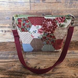 May include: A patchwork shoulder bag with a burgundy strap. The bag is made of hexagonal fabric pieces in various floral and patterned designs, including red roses, paisley, and gingham. The bag has a green floral lining.
