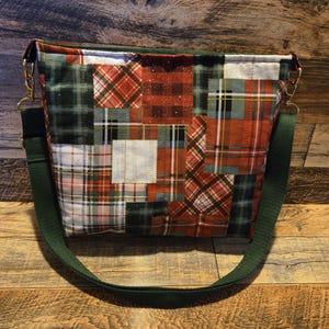 May include: A patchwork shoulder bag with a green strap and gold-colored hardware. The bag features a variety of plaid patterns in red, green, and white. The bag is sitting on a wooden surface.