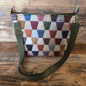 May include: A patchwork crossbody bag with a variety of fabric patterns in shades of blue, red, green, and brown. The bag has a green adjustable strap and metal hardware. The bag is on a wooden surface.
