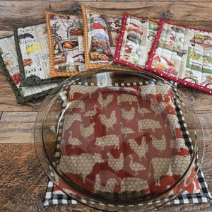 May include: A clear glass pie dish sits atop a red and white chicken-themed fabric. Surrounding it are several quilted pot holders with various farm-themed patterns, including eggs, vegetables, and farm animals. The pot holders have different colored borders.