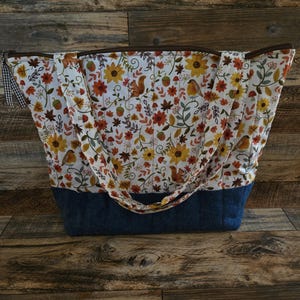 May include: A quilted tote bag featuring a white fabric upper section with a colorful autumn-themed print of sunflowers, leaves, acorns, squirrels, and birds. The lower section is solid dark blue fabric. It has two matching fabric shoulder straps.