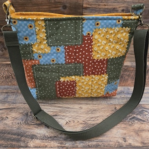 May include: A patchwork crossbody bag with a green adjustable strap. The bag features a variety of fabric squares in blue, yellow, brown, and green, with floral and polka dot patterns. The interior lining is yellow.