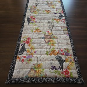 May include: A rectangular quilted table runner featuring a cream-colored fabric printed with black crows, orange jack-o'-lanterns, colorful flowers, and spider webs. The runner is finished with a black border patterned with white spider webs.