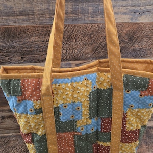 May include: A patchwork tote bag with a floral and polka dot pattern in shades of yellow, blue, green, and brown. The bag has mustard-colored straps and trim. The bag is made of quilted fabric.