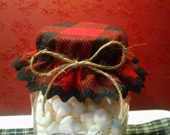 May include: A clear glass jar filled with white marshmallows, topped with a red and black plaid fabric lid. The lid is secured with twine tied in a bow. The jar sits on a white plate, with a red background.