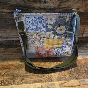 May include: A patchwork shoulder bag with a green strap. The bag is made of various floral and patterned fabrics in shades of blue, white, yellow, and red. The strap is adjustable and attached with silver-colored metal clips.