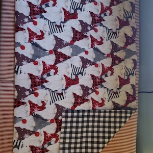 May include: A folded fabric with a repeating pattern of gnomes wearing red hats and white beards. The fabric is white with red, black, and gray accents. The reverse side is black and white checkered. A red and white striped fabric is visible on the left.