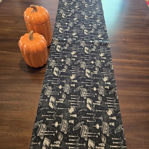 May include: A rectangular black table runner featuring a repeating pattern of white illustrations including open books, candelabras, ravens, and spiderwebs, accompanied by two orange ceramic pumpkin-shaped lidded jars with ribbed textures.