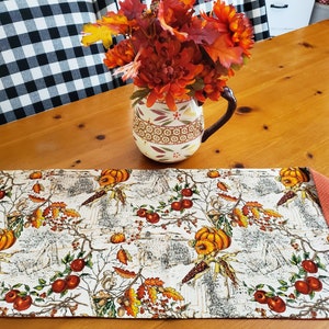 May include: A reversible table runner with a fall harvest theme. One side features a cream-colored fabric with pumpkins, apples, and corn stalks. The reverse side is a solid orange with white polka dots. A ceramic pitcher with fall foliage sits on the wooden table.