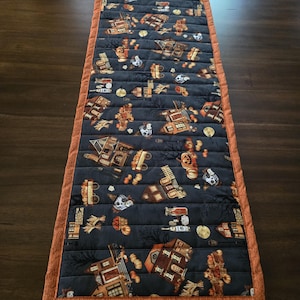 May include: A rectangular quilted table runner featuring a dark gray fabric printed with a pattern of houses, pumpkins, and skulls. The runner is finished with a solid orange fabric border and horizontal stitching lines across the entire surface.
