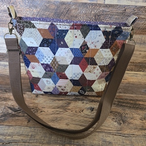 May include: A handmade patchwork tote bag with a brown adjustable strap. The bag features a geometric pattern of various fabric scraps in shades of brown, blue, red, and cream. The bag has a rectangular shape and is designed for carrying items.