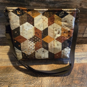 May include: A quilted crossbody bag with a geometric pattern of brown, tan, and cream-colored fabric. The bag features a long, adjustable strap and metal hardware. The patchwork design creates a 3D cube effect, giving the bag a unique, handcrafted look.