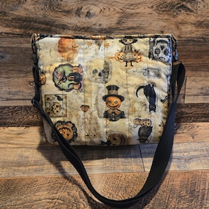 May include: A cream-colored Halloween-themed crossbody bag with a black strap. The bag features various spooky illustrations, including pumpkins, skulls, and black cats. The bag is rectangular and has a quilted appearance.