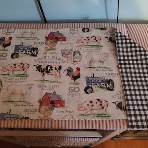 May include: A reversible farmhouse-themed fabric with a red and white striped border. One side features a cream background with farm animals and text, while the other side has a black and white checkered pattern.