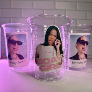 May include: Clear plastic cups featuring images of a woman, some with the text "Bday Girl" and "Birthday Girl". The woman is wearing sunglasses and holding a phone. The cups are on a white surface.