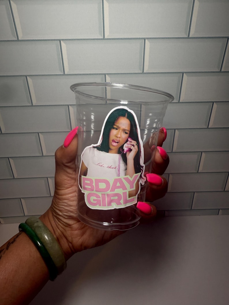 May include: A clear plastic cup with a sticker that says "BDAY GIRL" in pink and white. The sticker features a woman on the phone. The cup is held by a hand with bright pink nail polish and a green bracelet.