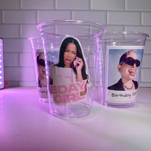 May include: Three clear plastic cups with images of women. One cup has the text "Bday Girl" and another has "Birthday Girl". The cups are on a white surface with a purple light.