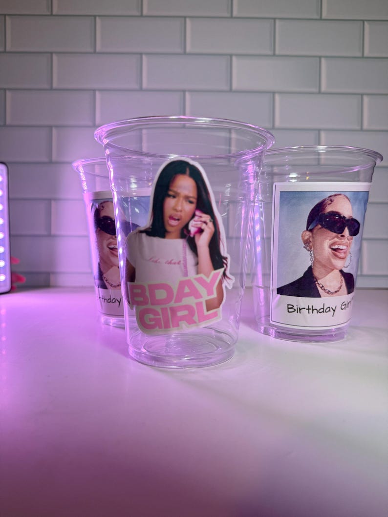 May include: Three clear plastic cups with images of women. One cup has the text "Bday Girl" and another has "Birthday Girl". The cups are on a white surface with a purple light.