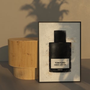 May include: A framed print featuring a black perfume bottle with the text "TOM FORD OMBRÉ LEATHER PARFUM" on a white background. The artwork is set against a neutral backdrop with a wooden side table.