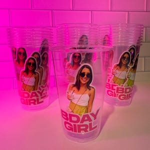 May include: Clear plastic cups featuring a photo of a person wearing sunglasses and the text "BDAY GIRL" in pink. The cups are stacked on a white surface, with a pink background.