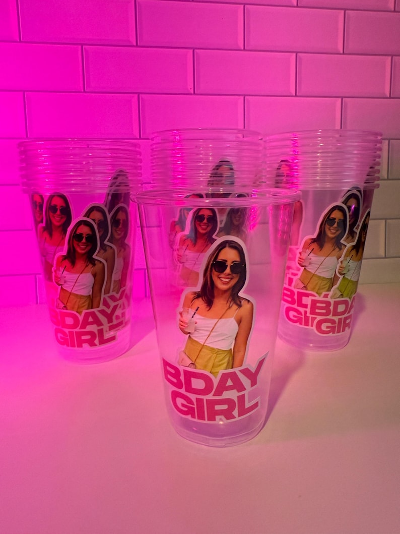May include: Clear plastic cups featuring a photo of a person wearing sunglasses and the text "BDAY GIRL" in pink. The cups are stacked on a white surface, with a pink background.