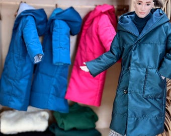May include: A doll wearing a teal hooded coat and plaid pants stands in front of a wooden wardrobe. The wardrobe contains blue and pink coats, sweaters, and three patterned caps on a shelf. The coats are hanging on hooks.