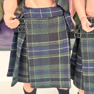 May include: A pleated kilt featuring a dark green, navy blue, and thin yellow plaid pattern. The garment includes two black fabric straps with silver-toned metal buckles on the side for adjustment, worn over black and green striped socks.