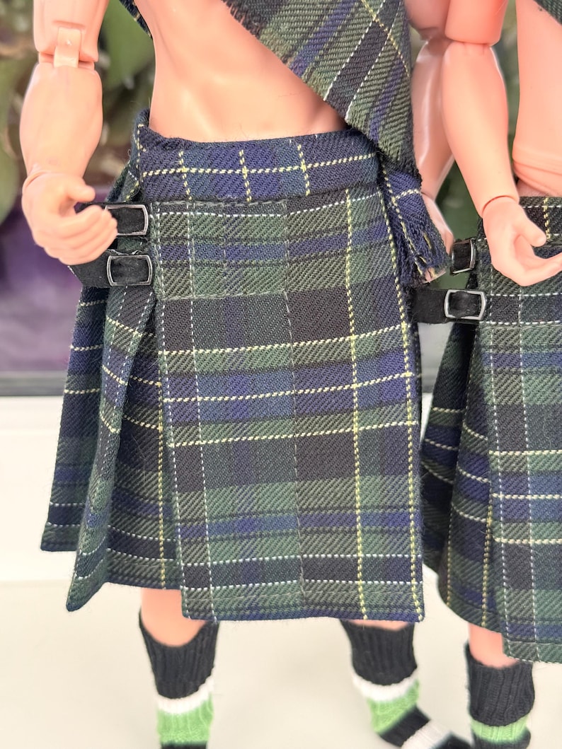 May include: A pleated kilt featuring a dark green, navy blue, and thin yellow plaid pattern. The garment includes two black fabric straps with silver-toned metal buckles on the side for adjustment, worn over black and green striped socks.