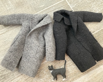 May include: Two miniature wool coats, one gray and one black, are displayed on a light wood surface. The coats have lapels and pockets. A small black and white cat figurine is placed near the coats.