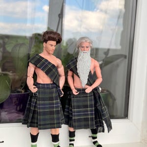 May include: Two male dolls wearing matching dark green, blue, and yellow plaid kilts and shoulder sashes. One doll has short brown hair, while the other has long gray hair and a long gray beard. Both dolls wear black, white, and green striped socks.