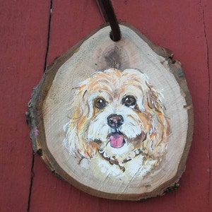May include: A hand-painted portrait of a small, light brown dog with a white muzzle and tongue sticking out, painted on a round wooden slice.