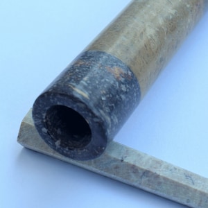 Op de afbeelding: Een lange, cilindrische marmeren pijp met een zwart en bruin marmerpatroon. De pijp rust op een kleiner, rechthoekig stuk marmer.