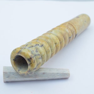 Op de afbeelding: Een lange, geel-bruine, gesneden stenen pijp met een ronde opening aan het ene uiteinde. De pijp rust op een kleiner, plat stuk steen.