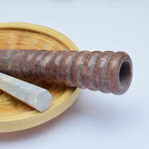 Può includere: Un mattarello di legno marrone con una superficie scanalata si trova su un cesto intrecciato. Una pietra rettangolare grigio chiaro si trova accanto al mattarello.