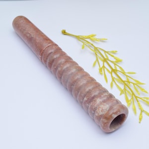 Può includere: Una pipa di pietra marrone, a spirale, con un'apertura liscia e arrotondata. La pipa è appoggiata su una superficie bianca con un ramo giallo e frondoso accanto.