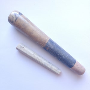 Può includere: Un set di due strumenti in pietra per macinare erbe e spezie. Lo strumento più grande è di forma cilindrica lunga con un motivo a marmo marrone e blu. Lo strumento più piccolo è di forma triangolare appuntita di colore grigio chiaro.