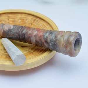 Könnte beinhalten: Eine braune und graue Steinpfeife mit einer runden Öffnung auf einem geflochtenen Bambus-Tablett. Ein weißer Stein befindet sich ebenfalls auf dem Tablett.