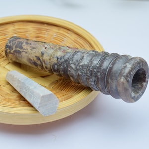 Könnte beinhalten: Eine braune und graue Steinpfeife mit einer runden Öffnung und einem kleineren, hellgrauen Stein. Die Pfeife liegt auf einem geflochtenen Korb.