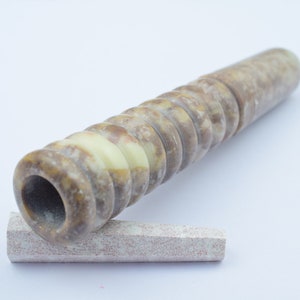 Könnte beinhalten: Eine braune und weiße Steinpfeife mit einer runden Öffnung und einem geriffelten Design. Die Pfeife liegt auf einem kleinen, rechteckigen Stück Stein.