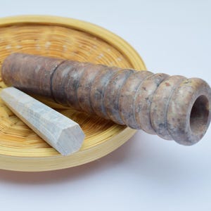 Op de afbeelding: Een set van een bruine en witte stenen mortier en vijzel. De mortier is een holle cilinder met een afgeronde bodem en een brede opening aan de bovenkant. De vijzel is een lang, cilindrisch stuk steen met een afgerond uiteinde. De set staat op een geweven mand.