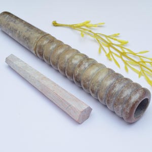 Op de afbeelding: Een bruine en groene stenen deegroller met een getextureerd oppervlak en een houten pen. De deegroller ligt op een wit oppervlak met een gele tak van kunstgroen.