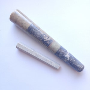 Könnte beinhalten: Ein Steinmörser- und Stößel-Set. Der Mörser ist zylindrisch mit einem Mosaikmuster aus Blau, Grau und Beige. Der Stößel ist ein kleiner, hellgrauer, sechseckiger Stab. Das Set befindet sich auf weißem Hintergrund.