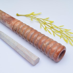 Op de afbeelding: Een bruine en witte marmeren deegroller met een getextureerd oppervlak en een witte houten pen. De deegroller ligt op een wit oppervlak met een groene tak met bladeren.